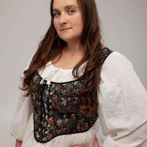 May include: A floral print corset with a black base, featuring a woodland design with foxes, rabbits, and birds. The corset has a lace-up front and is worn over a white blouse with long, puffy sleeves. The overall style is reminiscent of historical or renaissance fashion.