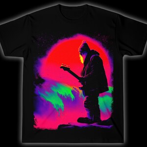 Puede incluir: Camiseta negra con una ilustración de neón de un guitarrista sobre un fondo colorido. El diseño incluye un sol rojo y salpicaduras de púrpura, verde y rosa. La persona está silueteada en negro, con gorro y abrigo.