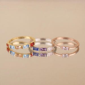 Op de afbeelding: Drie dunne metalen ringen op een rij. De linker ring is goudkleurig met drie baguette-geslepen stenen in rood, lichtblauw en oranje. De middelste ring is zilverkleurig met drie baguette-geslepen stenen in blauw, paars en roze. De rechter ring is roségoudkleurig met één roze steen.