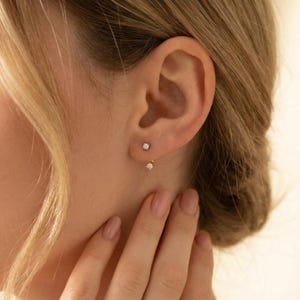 May include: A close-up of a person's ear adorned with two gold earrings. The top earring is a small, square stud. The second earring is a small, round diamond. The person's hand is visible, with manicured nails.