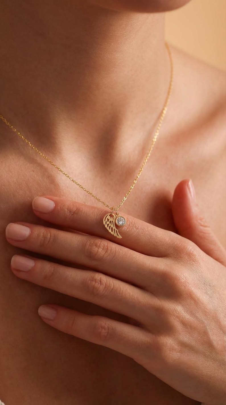 Pode incluir: Um delicado colar de ouro com um pingente de asa e uma pequena gema transparente e redonda. O pingente de asa tem detalhes intrincados. O colar &eacute; exibido na pele de uma pessoa.