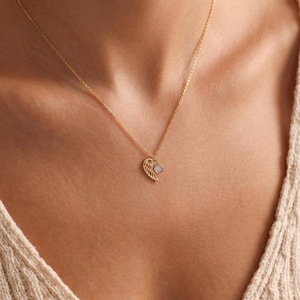 Pode incluir: Colar dourado com um pendente em forma de asa. O pendente de asa tem uma pequena pedra redonda e transparente. O colar &eacute; usado ao redor do pesco&ccedil;o, com um su&eacute;ter bege ao fundo.