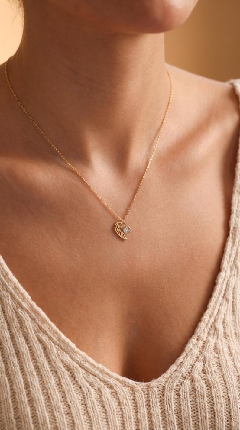 Pode incluir: Colar dourado com um pendente em forma de asa. O pendente de asa tem uma pequena pedra redonda e transparente. O colar &eacute; usado ao redor do pesco&ccedil;o, com um su&eacute;ter bege ao fundo.