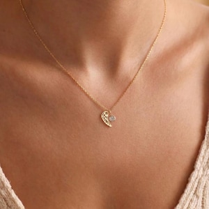 Pode incluir: Um colar dourado com um pingente delicado em forma de asa e uma pequena pedra preciosa transparente. O colar &eacute; exibido no peito de uma pessoa, com a corrente vis&iacute;vel. O pingente de asa tem detalhes intrincados.