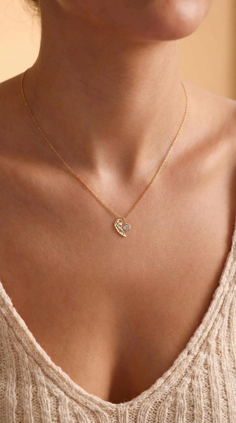 Pode incluir: Um colar dourado com um pingente delicado em forma de asa e uma pequena pedra preciosa transparente. O colar &eacute; exibido no peito de uma pessoa, com a corrente vis&iacute;vel. O pingente de asa tem detalhes intrincados.