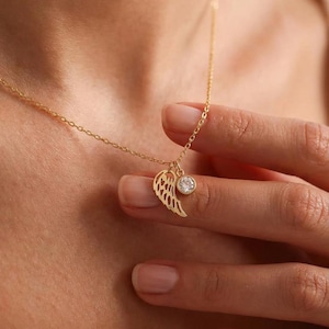 Pode incluir: Um delicado colar de ouro com um pingente de asa e um pequeno pingente redondo de pedra preciosa transparente. O colar &eacute; exibido em um fundo de tom de pele, destacando o design e a cor da joia.