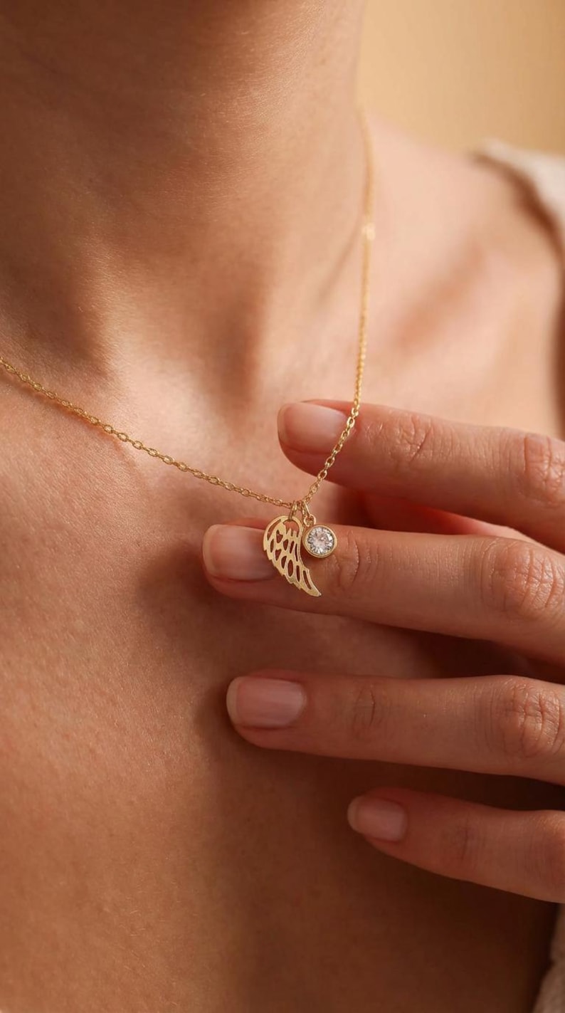 Pode incluir: Um delicado colar de ouro com um pingente de asa e um pequeno pingente redondo de pedra preciosa transparente. O colar &eacute; exibido em um fundo de tom de pele, destacando o design e a cor da joia.