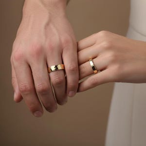 Puede incluir: Se muestran dos anillos de boda dorados, uno en cada mano. Los anillos tienen una superficie texturizada y un diseño sencillo y elegante. El fondo es de un tono neutro y cálido.