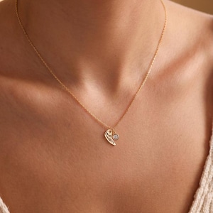 Pode incluir: Um colar dourado com uma corrente delicada. O pingente apresenta um design de asa e uma pequena pedra preciosa redonda e transparente. O colar &eacute; usado no pesco&ccedil;o de uma pessoa, contra uma blusa de cor clara.