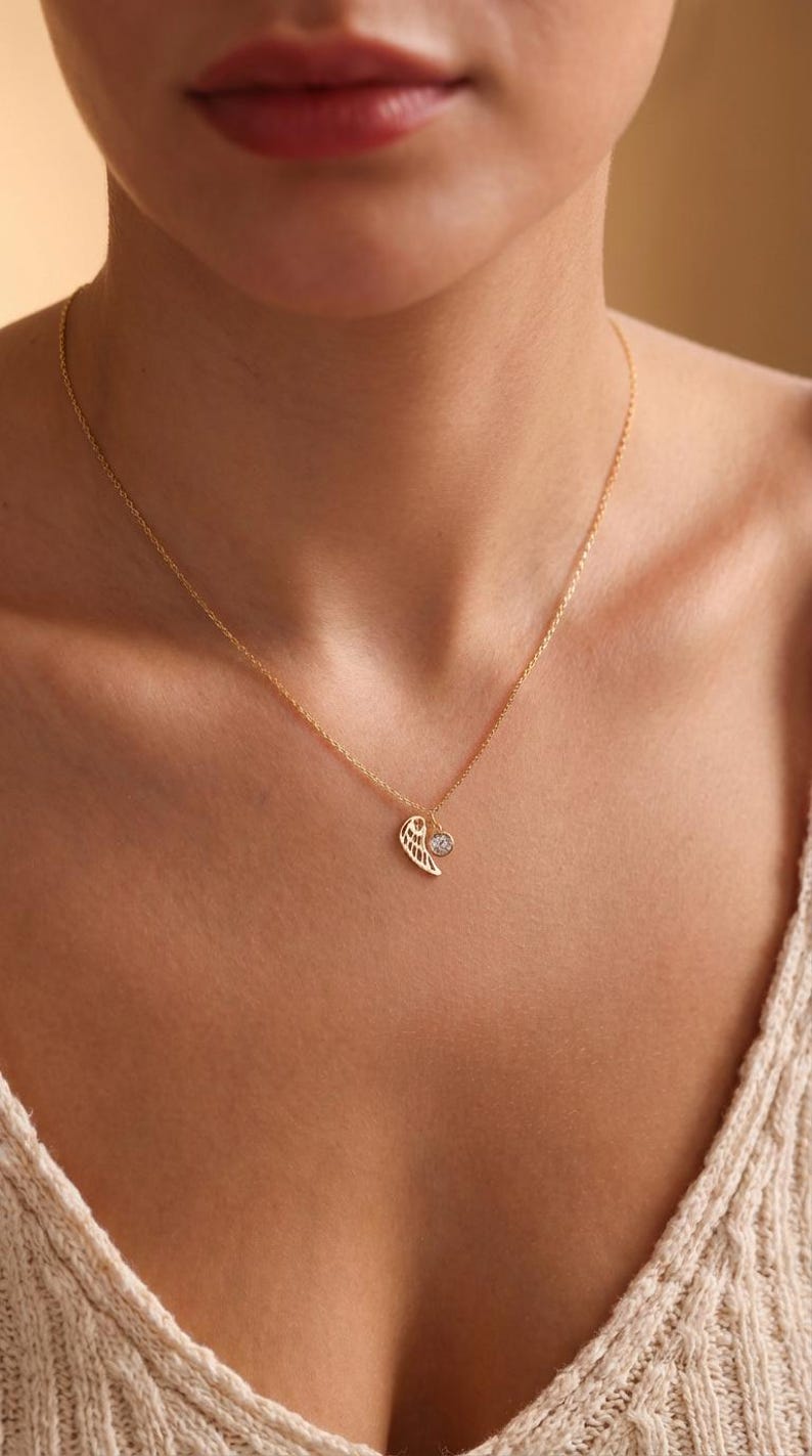 Pode incluir: Um colar dourado com uma corrente delicada. O pingente apresenta um design de asa e uma pequena pedra preciosa redonda e transparente. O colar &eacute; usado no pesco&ccedil;o de uma pessoa, contra uma blusa de cor clara.