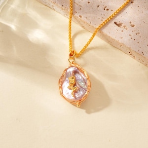 Op de afbeelding: Gouden ketting met een lavendelkleurige parel hanger. De hanger heeft een onregelmatige vorm, een gevlochten gouden draadframe en een kleine gouden rozenbedel. De ketting is een fijne gouden schakel.