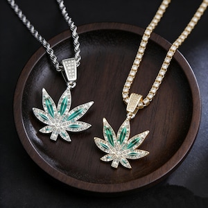 May include: Two necklaces featuring cannabis leaf pendants. One pendant is silver-toned with a rope chain, and the other is gold-toned with a tennis chain. Both pendants are inlaid with clear crystals and green enamel accents on the leaves.