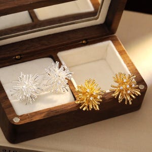 Pode incluir: Dois pares de brincos de pressão expostos numa caixa de joias de madeira. Um par é prateado e o outro dourado. Ambos têm um design espinhoso em forma de estrela com pequenas pedras transparentes.