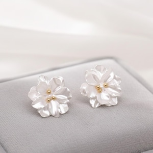 May include: A pair of white flower-shaped stud earrings. Each earring features five petals and three small gold-colored accents in the center. The earrings are displayed on a gray surface.