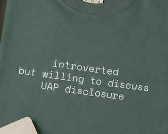 May include: A dark sage green crew neck t-shirt featuring the white text "introverted but willing to discuss UAP disclosure" printed on the chest. The shirt has a Comfort Colors brand label at the inner neckline and short sleeves.