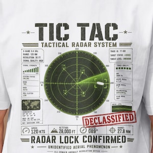 以下が含まれることがあります： TIC TAC TACTICAL RADAR SYSTEMと書かれた緑色の円形レーダー画面のグラフィックがプリントされた白いTシャツ。デザインには技術データ、赤いDECLASSIFIEDのスタンプ、RADAR LOCK CONFIRMED UNIDENTIFIED AERIAL PHENOMENONというテキストが含まれています。