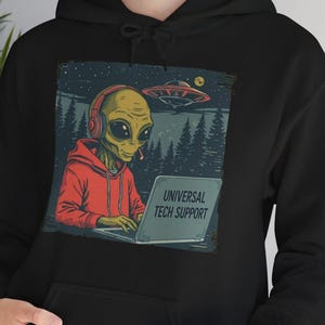 May include: Black hoodie with a graphic of an alien wearing headphones and a red hoodie, working on a laptop. The laptop displays the text "UNIVERSAL TECH SUPPORT". A UFO is in the background.