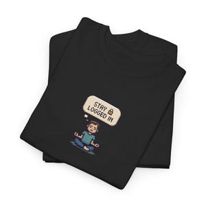 May include: Two black t-shirts are folded and stacked. The top shirt features a pixelated illustration of a person meditating with a thought bubble that reads "STAY LOGGED IN" with a lock icon. The person is wearing a blue shirt and blue pants.