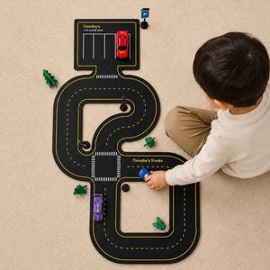 May include: A black modular toy road track set with yellow lane markings, featuring a parking area labelled Timothy's Play & Park Zone and a section marked Timothy's Tracks. Includes a red car, a purple car, a blue car, and green tree figures.