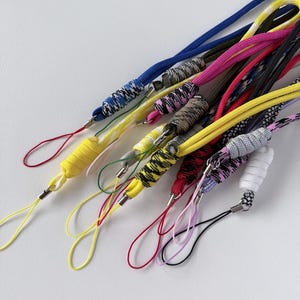 May include: A collection of colorful lanyards with various patterns and colors, including blue, pink, yellow, and black. Each lanyard has a loop at the end and a metal clasp. The lanyards are made of a woven cord.
