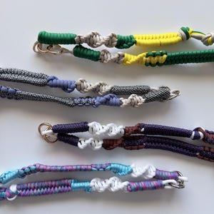 May include: Close-up of several handmade lanyards in various colors. The lanyards feature braided cord designs in green, yellow, blue, purple, and white, with metal clasp attachments. The cords are knotted in a decorative pattern.