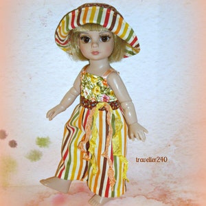 May include: A doll dressed in a striped jumpsuit and a matching hat. The jumpsuit has vertical stripes in brown, yellow, red, and green. The doll's top has a floral pattern. The hat has a wide brim with coordinating stripes. The doll is barefoot.