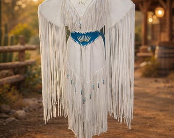 May include: A white fringed dress with a blue belt and turquoise accents. The dress has long fringe on the sleeves and hem, with decorative beadwork. The dress is displayed on a mannequin in an outdoor setting.