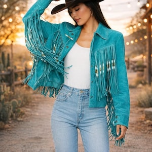 Puede incluir: Chaqueta de ante turquesa con largos flecos y detalles de cuentas. La chaqueta tiene cuello y cierre de botones. La modelo lleva un top blanco y vaqueros de lavado claro. La chaqueta es de estilo western.