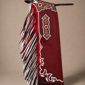 May include: A pair of deep red leather chaps featuring intricate floral tooling on the waistband and side panels. The design includes white maple leaf emblems, white leather piping accents, and long, layered fringe in red, white, and black.