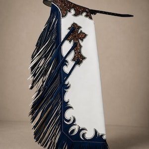 Peut inclure: Chaps en cuir blanc avec une ceinture et des bordures en cuir brun foncé à motifs floraux. Le panneau latéral est bleu marine avec de longues franges superposées bleu marine et blanches. Une croix en cuir brun est appliquée sur le panneau.
