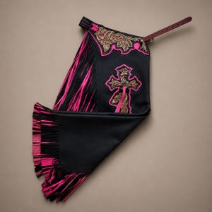 May include: Black leather chaps featuring vibrant hot pink fringe along the side and bottom edges. The upper section displays intricate tan floral tooling with hot pink trim and a prominent cross emblem, secured by a dark brown leather belt strap.