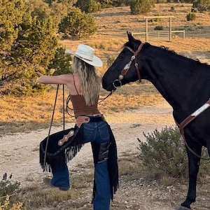 May include: A pair of black leather chaps with long fringe, worn over dark blue denim jeans, paired with a brown tank top and a white cowboy hat, standing next to a black horse with a brown leather bridle and silver-toned hardware accents.