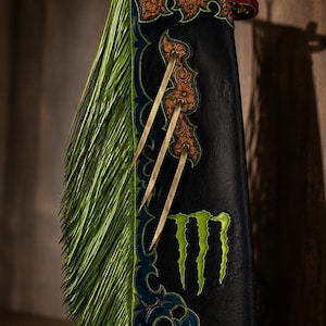 May include: Black leather chaps featuring long lime green fringe, intricate floral tooling at the top, and a blue decorative border. The side displays three tan leather ties and a bright green claw mark logo graphic printed on the lower section.