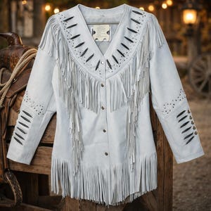 May include: A light blue suede jacket with long sleeves and a V-neck. The jacket features fringe detailing along the front, sleeves, and hem. It is adorned with black accents and small silver studs. The jacket is buttoned up and hangs on a wooden surface.