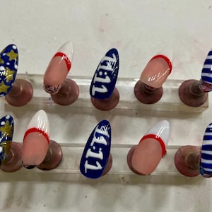 May include: A set of ten artificial nails with various designs. Some nails are navy blue with gold stars, others have a white tip with a red stripe, and some feature the number "11:11" in white on a navy blue background. One nail has blue and white stripes.