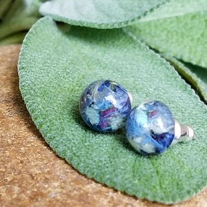 May include: Stud earrings featuring a clear resin design with blue, purple, and white floral inclusions. The earrings are set on silver posts. They are displayed on a green leaf.