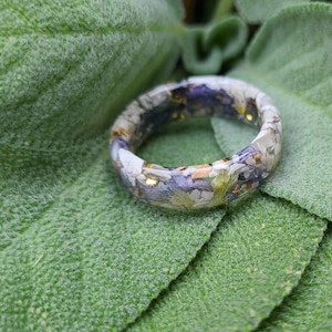 May include: A resin ring with a floral design. The ring features a mix of white, purple, and gold-colored elements. The ring is set against a green leaf background.