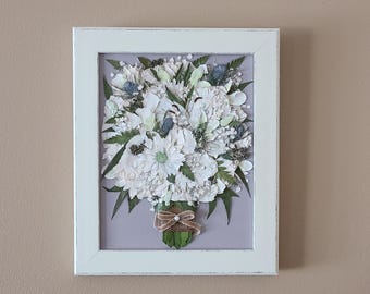 May include: A framed floral arrangement featuring a bouquet of white and green flowers. The bouquet includes daisies, and other flowers, with green leaves. The arrangement is set against a light purple background and framed in a white wooden frame.