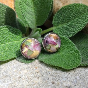 May include: A pair of round stud earrings featuring a silver-toned metal setting. The circular face of each earring contains a clear resin inlay embedded with small, colourful fragments of dried flower petals in shades of pink, green, and gold.
