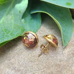 May include: A pair of round stud earrings featuring a gold-toned metal setting. The center of each earring is filled with a resin inlay containing small, multicolored fragments of dried flowers and metallic gold leaf flakes.
