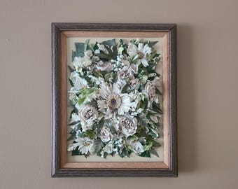May include: A framed floral arrangement featuring a variety of dried flowers in shades of white, cream, and muted rose. The arrangement includes daisies, roses, and greenery, all set against a light background within a wooden frame.