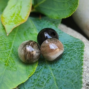 May include: Three round, decorative beads. Two are clear with hair-like inclusions, one brown and one with dark brown and light brown strands. The third bead is black with a hole. The beads rest on a green leaf.