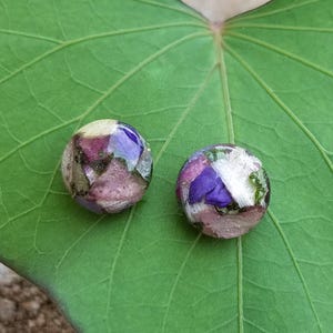 May include: A pair of round earrings, each encased in clear resin, featuring dried flowers in purple, pink, and white hues. They rest on a large, green leaf.