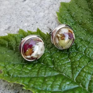 May include: A pair of round stud earrings featuring a polished silver-tone metal setting. Each earring contains a clear, domed resin cabochon encasing small, dried, multi-colored flower petals in shades of deep pink, white, and yellow.