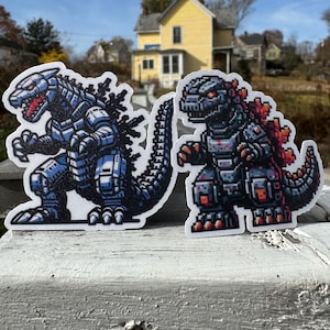 May include: Two pixelated Godzilla stickers. One is blue and silver, the other is black and red. Both have detailed designs and are placed on a weathered white surface with a blurred background of a yellow house and trees.
