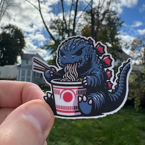 May include: A sticker featuring a blue cartoon monster eating noodles from a cup. The monster has red dorsal fins and is holding chopsticks. The cup has a red circle design. The sticker is held in front of a green lawn and a cloudy sky.