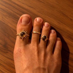 Puede incluir: Tres anillos para los dedos del pie en tono dorado. El primer anillo presenta un diseño de rayos de sol con una piedra oscura central. El segundo es una banda lisa y sencilla. El tercer anillo es una banda simple con una pequeña piedra oscura ovalada.