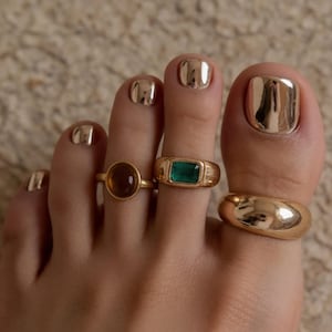 Può includere: Tre anelli color oro indossati sulle dita dei piedi. Un anello presenta una pietra rotonda color ambra, un altro mostra una pietra rettangolare verde smeraldo e il terzo è una fascia color oro lucido. Le unghie dei piedi hanno una finitura oro metallizzato.