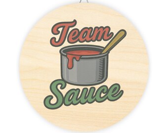May include: Circular wooden sign featuring the text "Team Sauce" in red and green. A grey pot filled with red sauce and a wooden spoon is in the centre. The sign has a metal hanger at the top.