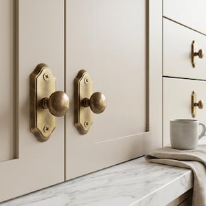 Peut inclure: Gros plan sur une armoire de cuisine avec des ferrures en laiton. Les portes et les tiroirs sont de couleur beige clair. Une tasse grise et un chiffon beige plié sont posés sur le comptoir. La quincaillerie comprend des boutons et des poignées.